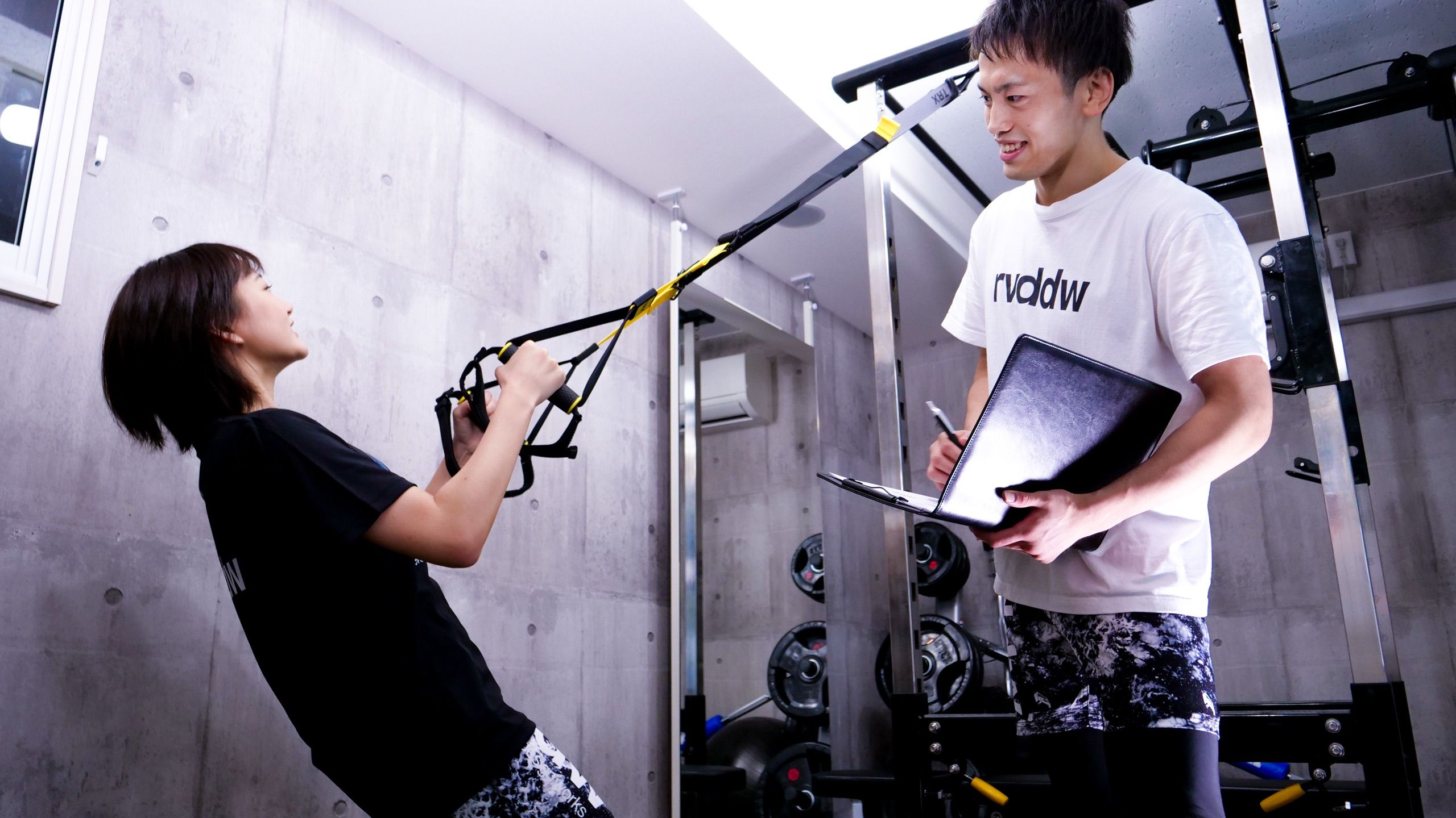 FUMAKE GYM 西新宿の完全個室で、TRXを使うお客様をトレーナーがメモを取りながら指導する様子